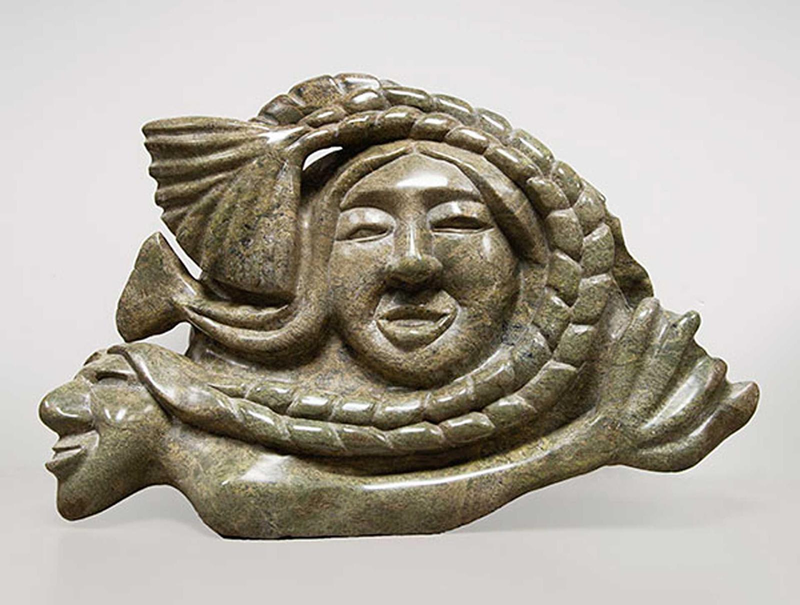 Green stone carving of a woman's face surrounded by a fish tail braid, supported by a prone figure of a man with a fish tail.