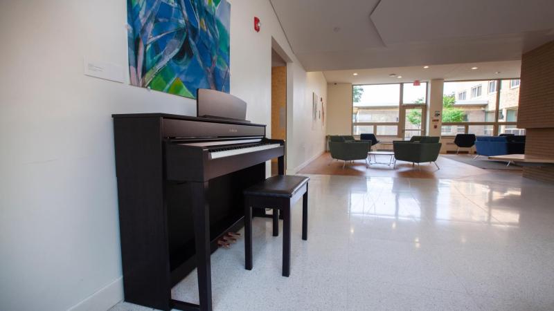 black upright piano against white wall with decorative poster in dorm common room, green lounge chairs and large windows in background